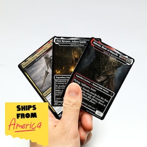 Op de afbeelding: Drie verzamelkaarten worden in een hand gehouden. De kaarten bevatten fantasy-kunst en tekst, waaronder "Nameless King, Storm Heir", "Fire Keeper, Ashen Guide" en "Malenia, Blade of Miquella". De kaarten zijn zwart met kleurrijke illustraties.