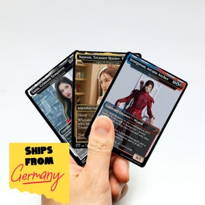 May include: Three collectible trading cards featuring images of women. The cards are titled "Sana, Crimson Temptress", "Nayeon, Treasure Hunter", and "Jeongyeon, Arcane Archer". The cards are held in a hand against a white background.