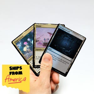 May include: Three Magic: The Gathering trading cards are held in a hand. The cards feature fantasy art and text, including "Magical Forest," "Utopia," and "Uncharted Self." A yellow square with the text "SHIPS FROM America" is in the lower left.
