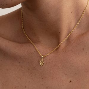 Può includere: Una collana color oro con un ciondolo a forma di mano di Hamsa. Il ciondolo è un piccolo ciondolo dettagliato. La catena è un delicato stile a corda ritorta. La collana è indossata intorno al collo.
