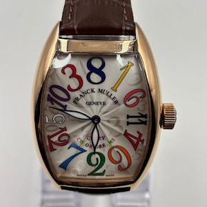 Könnte beinhalten: Eine goldfarbene Armbanduhr mit braunem Lederarmband. Das Zifferblatt der Uhr ist silberfarben mit bunten Ziffern und den Worten "FRANCK MULLER GENEVE" und "COLOR DREAMS". Die Uhr hat eine geschwungene rechteckige Form.