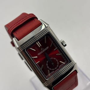 Könnte beinhalten: Eine rechteckige Armbanduhr mit rotem Zifferblatt und rotem Lederarmband. Die Uhr hat ein silberfarbenes Gehäuse und die Marke "Jaeger-LeCoultre" ist sichtbar. Sie hat ein kleines Sekunden-Zifferblatt.