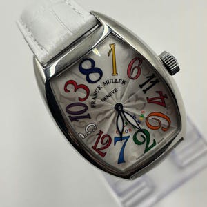Könnte beinhalten: Eine silberne Franck Muller Geneve Uhr mit weißem Lederarmband. Das Zifferblatt hat ein silbernes Radialmuster mit bunten Ziffern. Die Uhr hat eine Krone und die Worte "COLOR DREAMS" sind sichtbar.