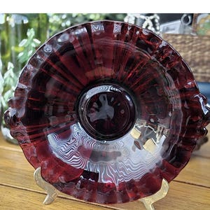 May include: A deep red glass bowl with a ruffled edge, resting on a gold-coloured metal stand. The bowl has a circular centre and a textured pattern. The stand has three legs.