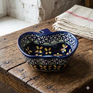 May include: A blue ceramic apple-shaped bowl with a decorative floral pattern. The bowl features a dark blue exterior with white, yellow, and green floral designs. The bowl sits on a rustic wooden surface.