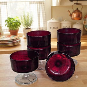 May include: A set of five deep red glass dessert bowls, each featuring a wide, circular bowl atop a clear, thick glass pedestal base. The bowls have a smooth, translucent finish and are arranged in a cluster on a light-colored wooden surface.