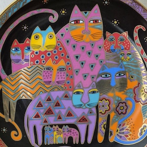May include: A decorative ceramic plate featuring a black background with a colorful illustration of multiple cats. The cats are depicted in various shades of pink, orange, blue, and purple, adorned with geometric patterns, dots, and floral motifs.