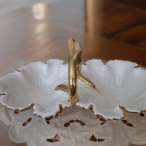May include: A white, two-sectioned serving dish with a gold handle. The dish has a scalloped edge with gold trim. It sits on a white, lace-edged doily. The handle is a stylized gold design.