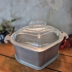 May include: A vintage, square-shaped casserole dish with a clear glass lid. The dish is made of silver-colored metal with a textured surface and two handles. The lid has a decorative handle and etched designs. The dish sits on a wooden surface.