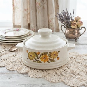 May include: A cream-coloured ceramic casserole dish with a lid, featuring a speckled finish and a decorative band of yellow and orange flowers with green leaves. The dish has two side handles and sits on a white lace doily next to a silver spoon.