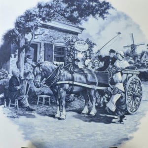 May include: A round ceramic plate featuring a blue and white transferware scene of a horse-drawn carriage in front of a brick building. The illustration depicts a person playing an accordion, a horse, and people in traditional clothing near a windmill.