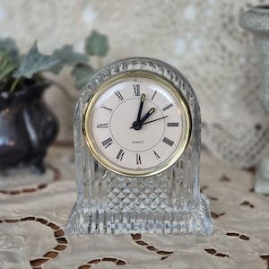 May include: A clear quartz clock with a gold-tone bezel and Roman numeral hour markers. The clock has a textured, crystal-like appearance and a rectangular base. The clock face reads "QUARTZ".
