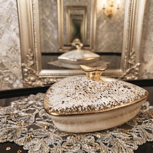 May include: A heart-shaped ceramic trinket box with a cream-colored base and a lid featuring a speckled brown and gold finish. The lid is topped with a small, gold-toned handle shaped like a crescent, and the edges are trimmed in metallic gold.