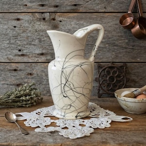 May include: A cream-colored ceramic pitcher featuring a black splattered paint design. The pitcher has a curved handle, a wide pouring spout, and a flared base. The surface is smooth and glossy, showcasing thin, irregular black lines.