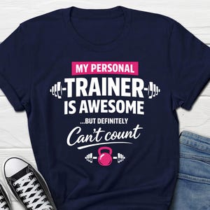Könnte beinhalten: Marineblaues T-Shirt mit dem Text "MY PERSONAL TRAINER IS AWESOME...BUT DEFINITELY Can't count". Das Design umfasst weißen Text und eine rosa Kettlebell-Grafik. Weiße Turnschuhe und blaue Jeans sind sichtbar.