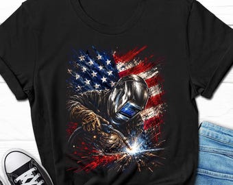 May include: A black t-shirt featuring a graphic print of a welder wearing a protective helmet and gloves, holding a welding torch that emits bright sparks. The background of the graphic displays a stylized, distressed American flag in red, white, and blue.