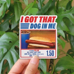 May include: A sticker with the text "I GOT THAT DOG IN ME" in red and blue. The sticker features an image of a hot dog in a bun, a blue soda cup, and the text "1/4 POUND PLUS ALL BEEF HOT DOG AND 20 oz. SODA".