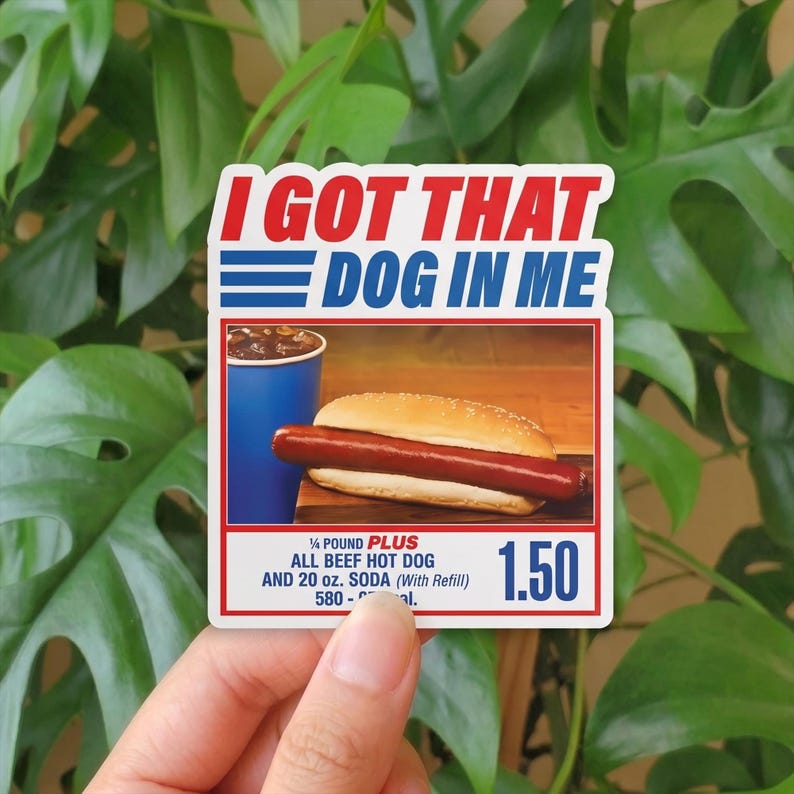 May include: A sticker with the text "I GOT THAT DOG IN ME" in red and blue. The sticker features an image of a hot dog in a bun, a blue soda cup, and the text "1/4 POUND PLUS ALL BEEF HOT DOG AND 20 oz. SODA".