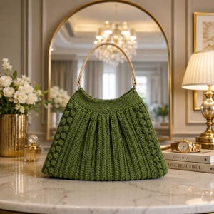 May include: A sage green crochet handbag featuring a structured pleated design, vertical textured bobble stitch columns on the sides, and a single light beige leather-like handle attached with silver-toned metal hardware rings.