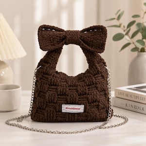 May include: A dark brown crocheted handbag featuring a basket-weave texture, a large bow accent at the top, and a silver-toned metal chain strap. A rectangular white fabric label with the red text Providence is stitched to the front centre.