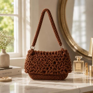 May include: A rust-colored handbag crafted with a textured, crocheted pattern. The bag features a single, thick, braided shoulder strap attached to the main body with two silver-toned metal rings. The base is constructed with a dense, solid stitch.