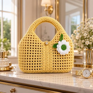 Può includere: Una borsa gialla all'uncinetto con un abbellimento floreale. La borsa presenta un design traforato e un manico arrotondato. Un fiore bianco e verde è attaccato sul davanti. La borsa è su una superficie di marmo.