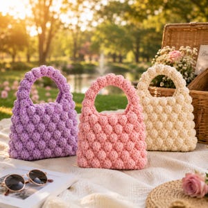 May include: Three small handbags made of crocheted yarn, each featuring a textured bobble stitch pattern. The bags are arranged in a row and come in three distinct colors: a light purple, a soft pink, and a creamy off-white shade.