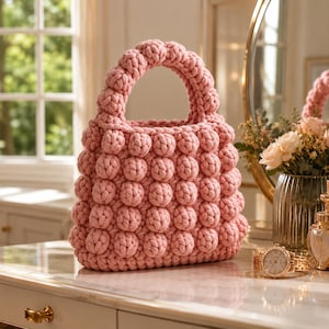 Pode incluir: Uma mala de mão rosa claro feita com um padrão de pontos de bolha texturizados em todo o corpo e na alça. A mala apresenta uma forma arredondada com uma construção de malha grossa, exibindo uma série uniforme de nós de fio elevados.