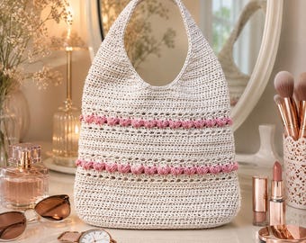 Könnte beinhalten: Eine cremefarbene Häkeltasche im Hobo-Stil mit einem einzelnen integrierten Tragegriff. Die Tasche ist mit zwei horizontalen Bändern aus erhabenen, strukturierten rosa Noppenstichen verziert, die über den gewebten Körper verlaufen.