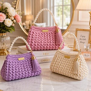 May include: Three crocheted handbags in pink, lavender, and cream. Each bag features a white pearl bead handle, a gold-toned rectangular brand plate, and a matching leather-like tassel attached to a gold-toned metal clasp on the side.