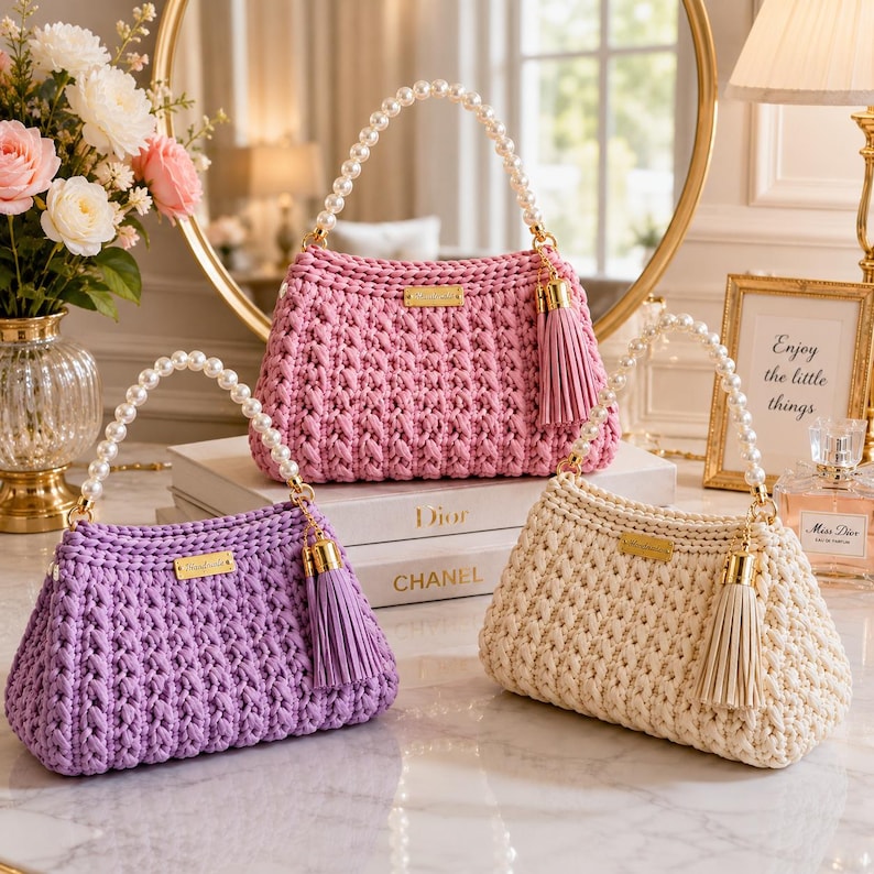 May include: Three crocheted handbags in pink, lavender, and cream. Each bag features a white pearl bead handle, a gold-toned rectangular brand plate, and a matching leather-like tassel attached to a gold-toned metal clasp on the side.