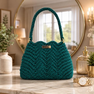 May include: A dark teal crochet handbag featuring a chevron pattern, a single rounded handle, and a small rectangular gold-toned metal brand plate centered on the front. The bag is constructed with a textured yarn stitch and gold-toned hardware.