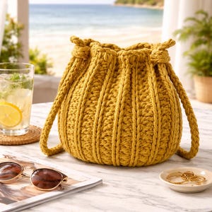 Puede incluir: Bolso de crochet amarillo mostaza con asas trenzadas. El bolso tiene un cierre de cordón y un diseño acanalado texturizado. El bolso está sobre una mesa de mármol con una revista, gafas de sol y un plato pequeño con joyas.