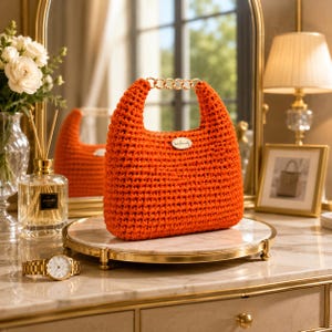 May include: A bright orange crochet handbag featuring a textured knit pattern, a gold-toned chain link handle, and a small oval-shaped tag centered on the front with the word handmade written in cursive script.