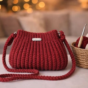 Può includere: Una borsa all'uncinetto rossa fatta a mano con una lunga tracolla. La borsa ha una trama a coste e una piccola etichetta metallica rettangolare. Un cestino con filato e un uncinetto è sullo sfondo.