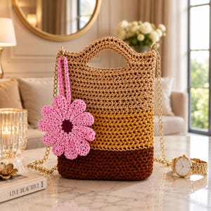 May include: A crocheted tote bag with a handle and gold chain straps. The bag features a color gradient from light tan to brown, with a pink flower charm. A gold watch with a gold band is next to the bag.