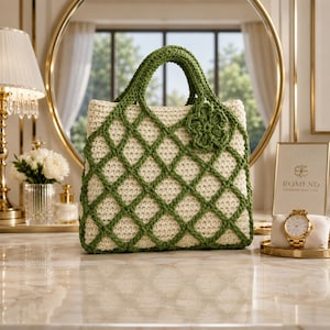 May include: A cream-colored crochet tote bag with a green diamond pattern and matching green handles. A green flower-shaped embellishment is attached to the front. The bag is sitting on a marble surface.