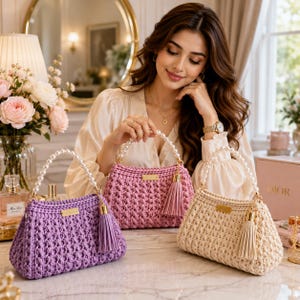 May include: Three handmade crochet handbags in purple, pink, and cream. Each bag features a textured knit pattern, a gold-tone metal brand tag, a matching leather-look tassel, and a handle made of white faux pearls with gold-tone hardware.