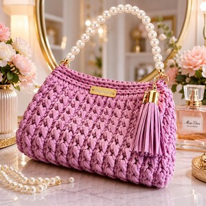 May include: A lavender crochet handbag featuring a thick, textured knit pattern, a handle made of white spherical pearls, and a matching lavender leather-like tassel attached to a gold-toned metal ring. A small rectangular gold-toned metal plaque is centered.
