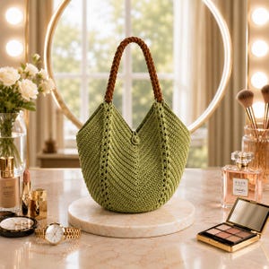 Può includere: Una borsa all'uncinetto color verde salvia con un motivo a chevron a forma di V, un unico manico intrecciato marrone e una piccola chiusura centrale a bottone. La borsa ha una base arrotondata e una silhouette strutturata con dettagli traforati.
