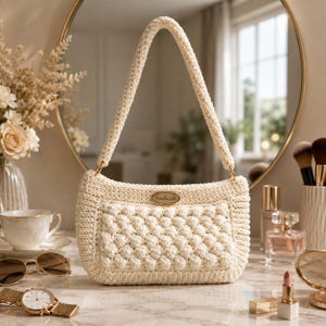 May include: A cream-colored crochet handbag featuring a textured bobble stitch pattern on the front panel, a single shoulder strap, and a small oval metal tag with the word handmade engraved in the center, attached to the front of the bag.