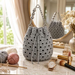 Puede incluir: Un bolso gris tejido a ganchillo con cordón y asa de cadena plateada y cuentas. El bolso tiene un diseño calado. El fondo incluye ovillos de lana, un reloj y artículos de maquillaje, lo que sugiere un accesorio hecho a mano.