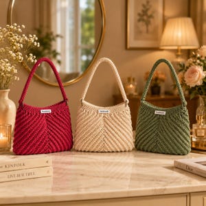 Puede incluir: Tres bolsos de ganchillo en rojo oscuro, crema y verde salvia. Cada bolso presenta una textura de punto con patrón de chevrón, una sola correa para el hombro y una pequeña etiqueta rectangular blanca en el centro con la palabra handmade impresa.