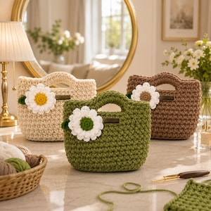 May include: Three crochet handbags in cream, olive green, and brown, each adorned with a large daisy. The bags have a rectangular shape with a handle and a small tag. The daisy has white petals and a yellow or brown center.