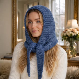 Könnte beinhalten: Eine blaue Strickhaube mit langen Bindebändern, getragen über einem weißen Pullover. Die Haube ist aus strukturiertem Garn gefertigt und bedeckt Kopf und Hals, mit Bändern, die unter dem Kinn gebunden werden können. Der Hintergrund zeigt einen Spiegel und Blumen.