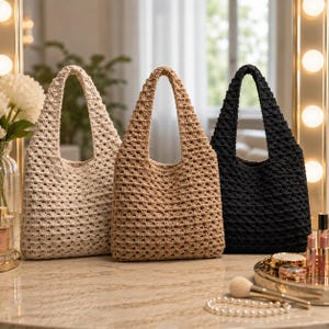 May include: Three crochet tote bags in cream, tan, and black colors. Each bag features a consistent open-weave pattern, a single integrated handle, and a square-shaped body. The bags are arranged side-by-side, showcasing their textured construction.