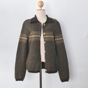 May include: A brown, button-down cardigan with a geometric pattern. The sweater has a dark brown collar and cuffs, with horizontal stripes of white and gold near the chest. The cardigan is displayed on a mannequin.