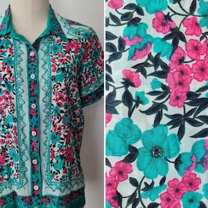 May include: A short-sleeved button-down shirt with a floral pattern. The shirt features a white background with teal and pink flowers, and teal trim. The shirt is displayed on a mannequin.