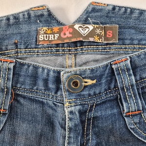 May include: Close-up of blue denim jeans with orange stitching and a button closure. A brown tag with the text "SURF &" and a heart logo is visible. The jeans are labeled size "S".
