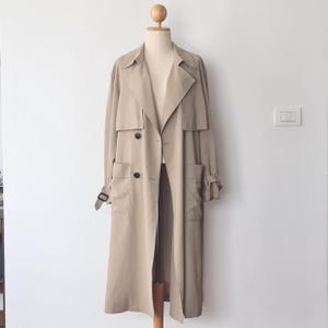 以下が含まれることがあります： マネキンに展示されたベージュのトレンチコート。コートは、大きなボタンが付いたダブルブレストフロント、ノッチカラー、ポケットが特徴です。長いコートにはベルトと調節可能な袖口が付いています。この衣服は軽量の生地で作られています。